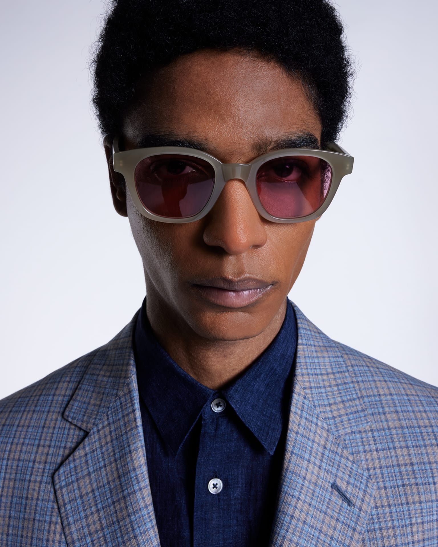 a headshot of a male model wearing a blue checked suit jacket and navy shirt, accessorising with light brown sunglasses with red tinted lenses