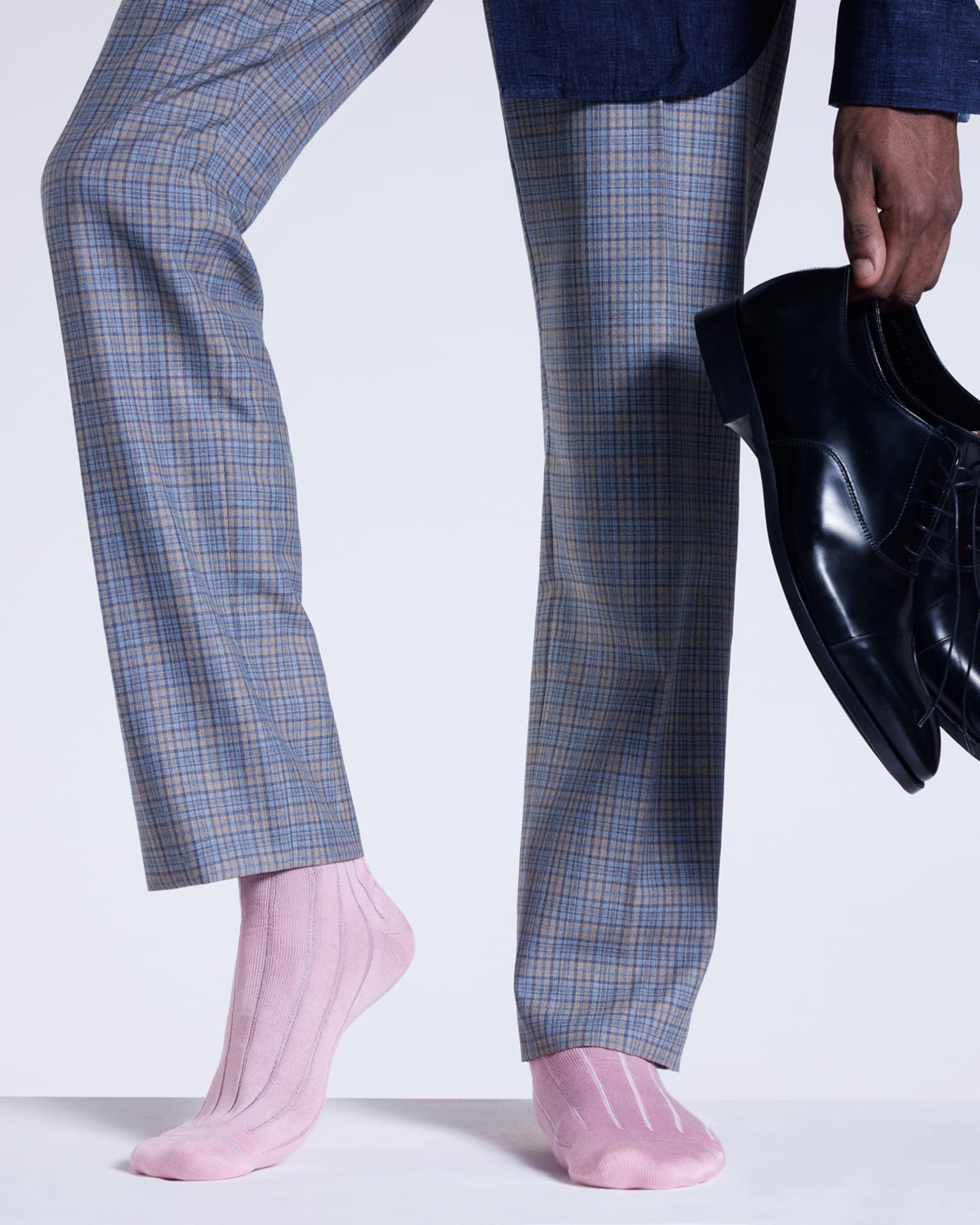 a male models legs wearing pink socks, checked trousers and a blue shirt holding a pair of black oxford shoes