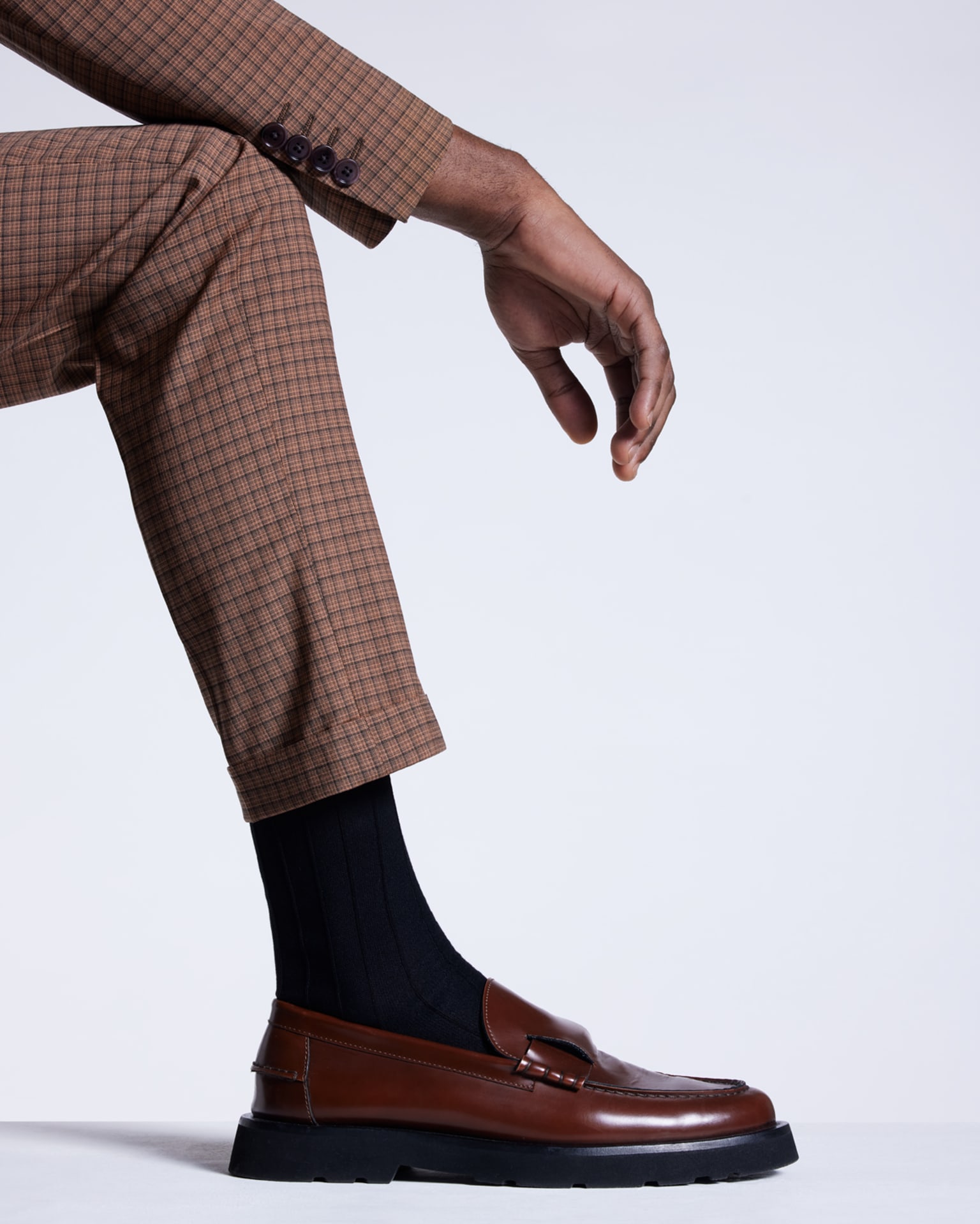 a male model resting his arm on his leg wearing brown checked suit trousers, black socks and brown loafers