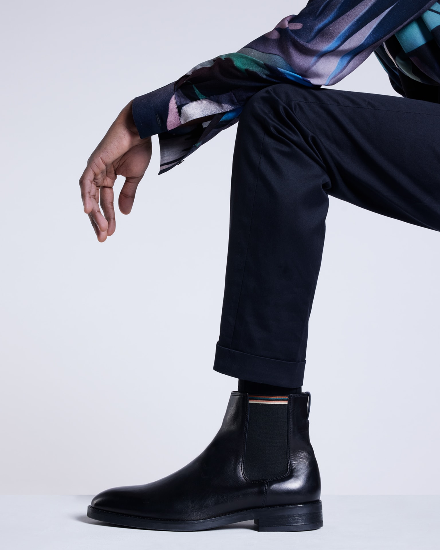 a male model resting his arm on his leg wearing a blue patterned statement shirt, navy trousers and black leather chelsea boots