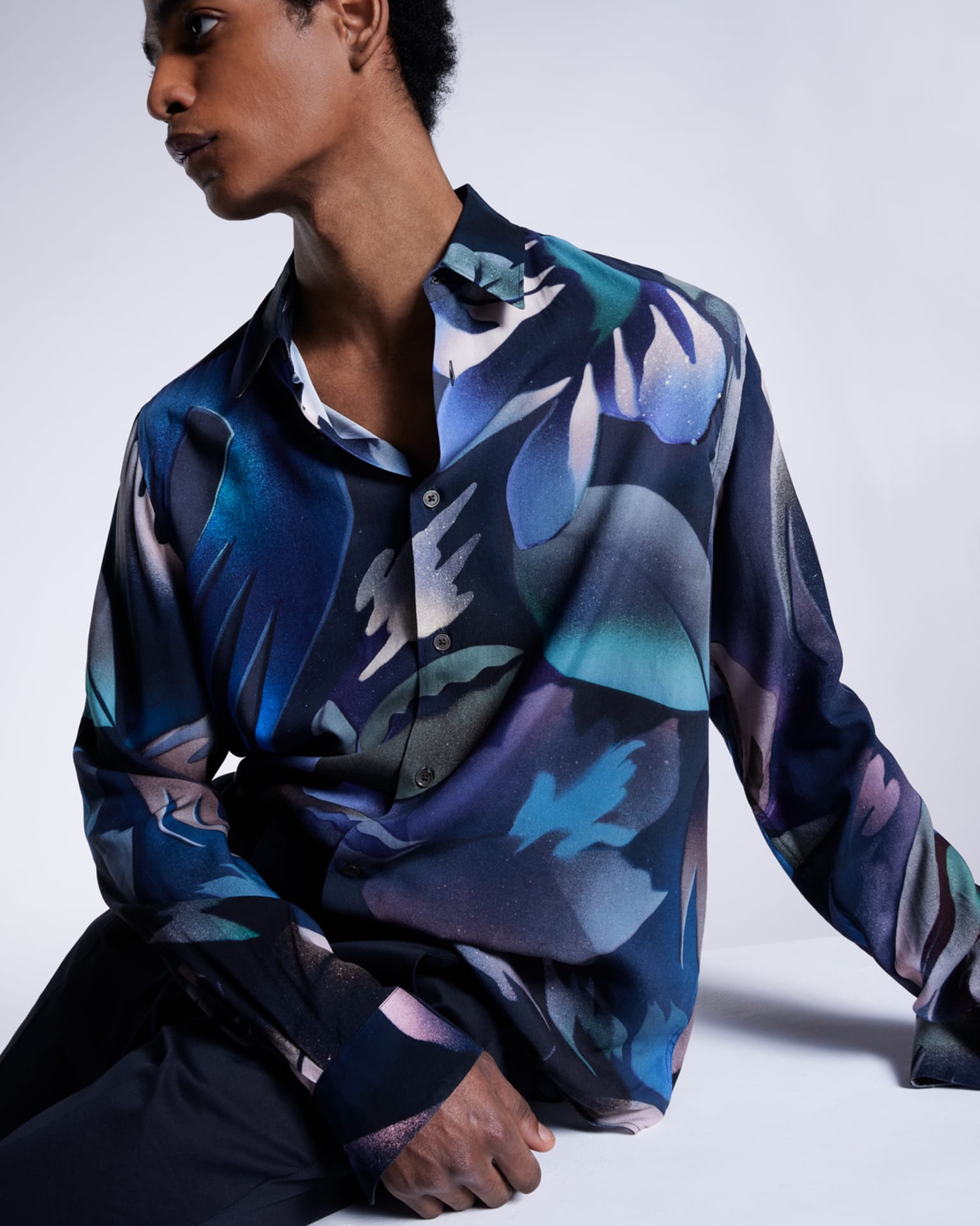 a male model sat on a white box wearing a colourful patterned statement shirt in blue, purple and green tones