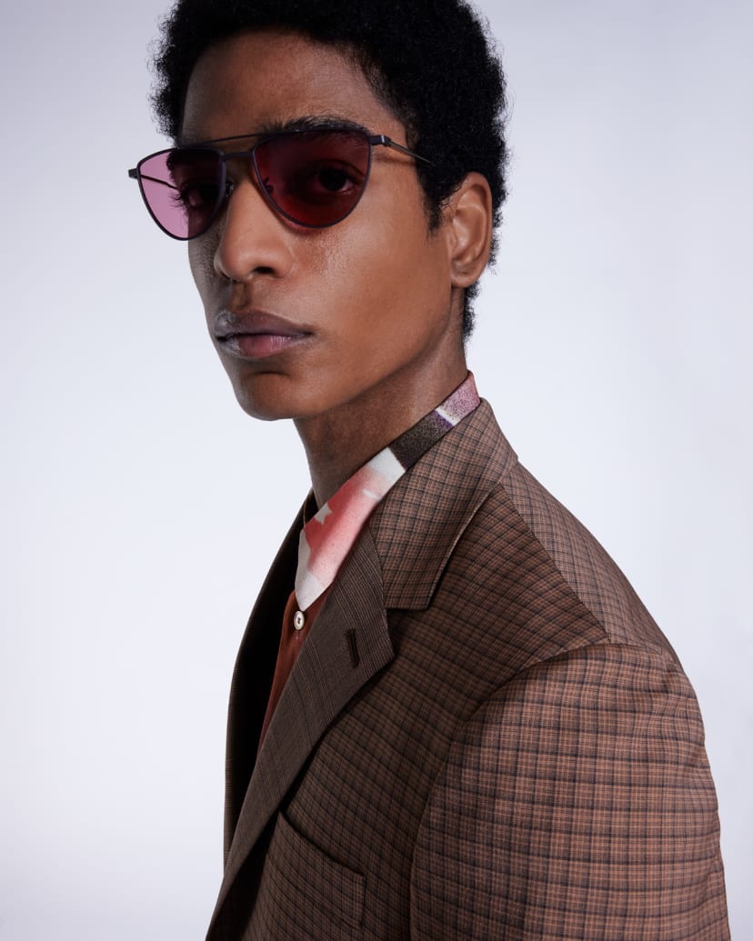 a male model wearing a brown checked suit jacket, statement shirt and sunglasses with a black frame and red tinted lenses