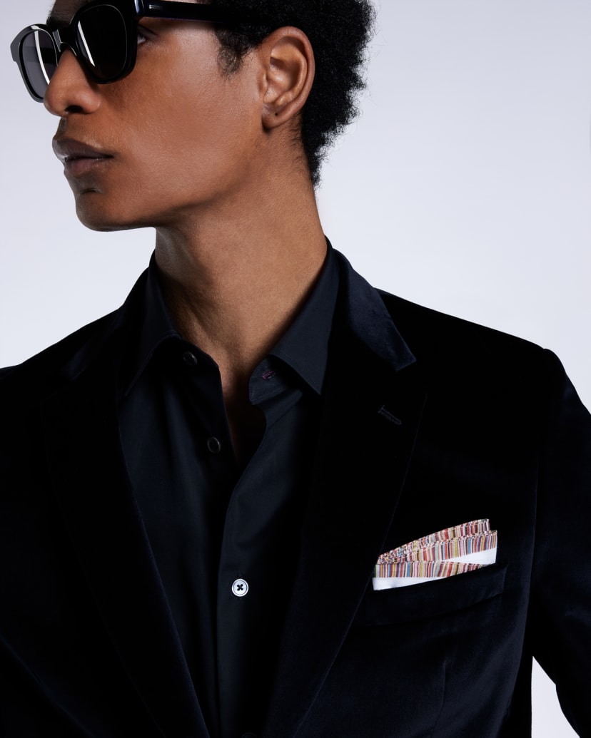 a male model wearing a black shirt and velvet suit jacket, paired with a colourful striped pocket square and black sunglasses